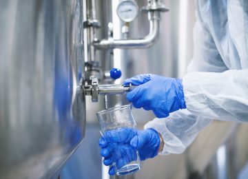 Close up view of hands in protective gloves opening tap valve and filling glass with samples from industrial machine reservoir.